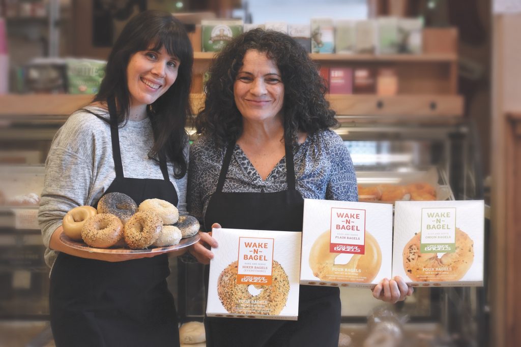 Francesca and Angelina, smiling with a tray of organic bagels and packaged products at Little Shop of Bagels bakery.