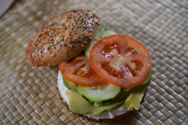 A freshly prepared organic bagel sandwich with layers of cream cheese, cucumber, and tomato on a woven mat from Little Shop of Bagels.
